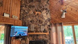 Un interior rústico con pared de piedra, vigas de madera, una cabeza de venado montada, chimenea y un televisor mostrando una competición de carreras.