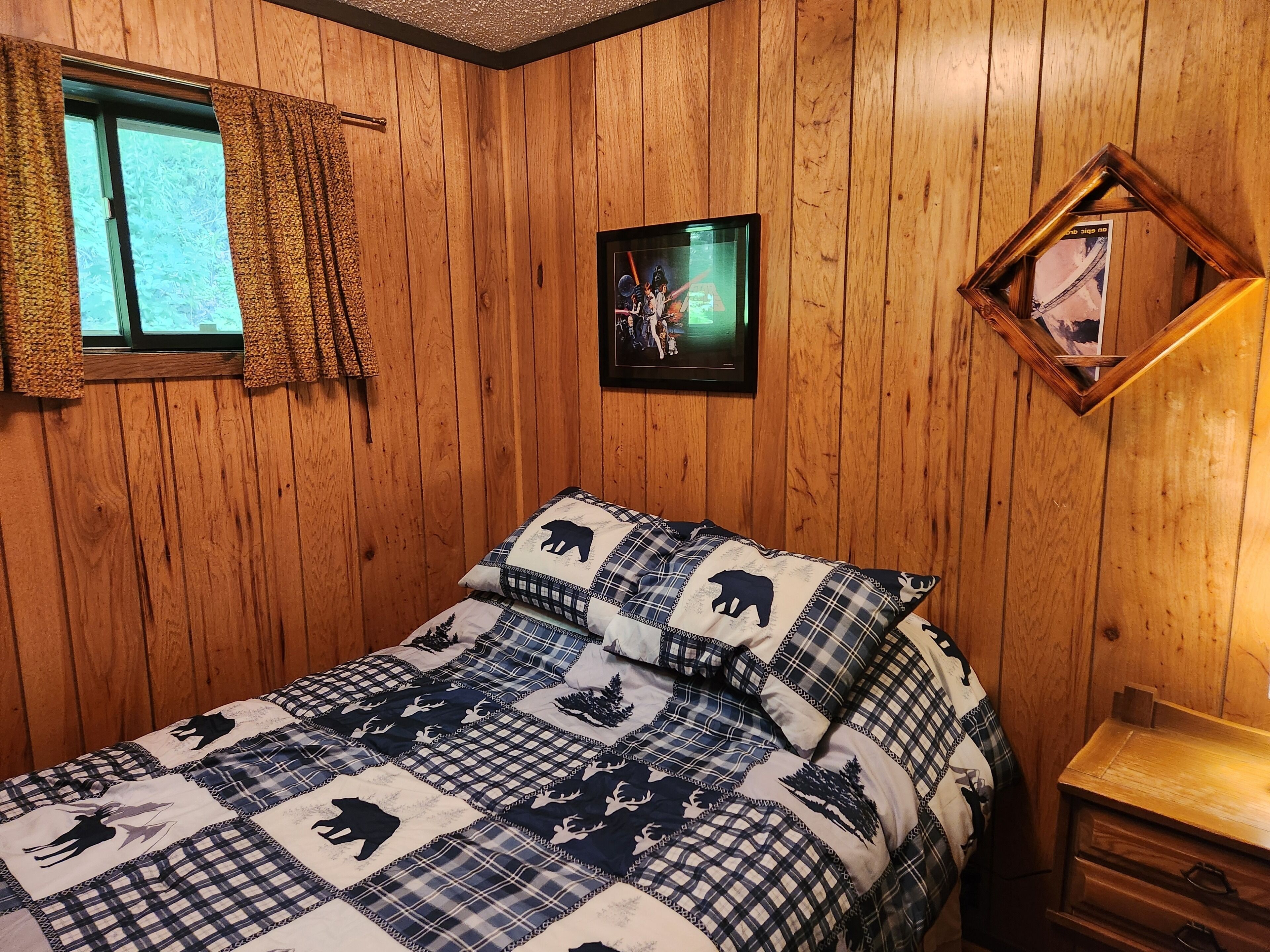 Ein Zimmer mit Holzwandverkleidung, einem Bett mit kariertem Quilz, einem Fenster mit Vorhängen, einem gerahmten Bild an der Wand und einem Nachttisch aus Holz.