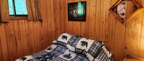Ein Zimmer mit Holzwandverkleidung, einem Bett mit kariertem Quilz, einem Fenster mit Vorhängen, einem gerahmten Bild an der Wand und einem Nachttisch aus Holz.