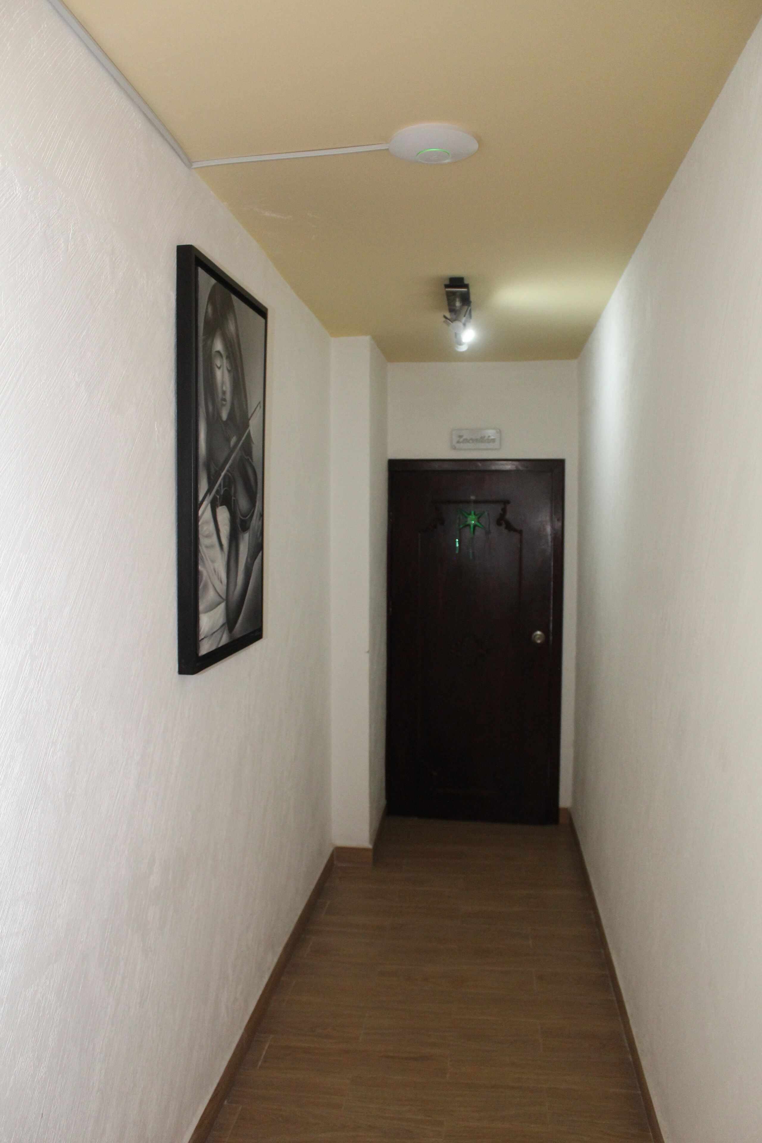 A narrow hallway with a wooden floor, a black door with a green sign, and a ceiling light fixture.