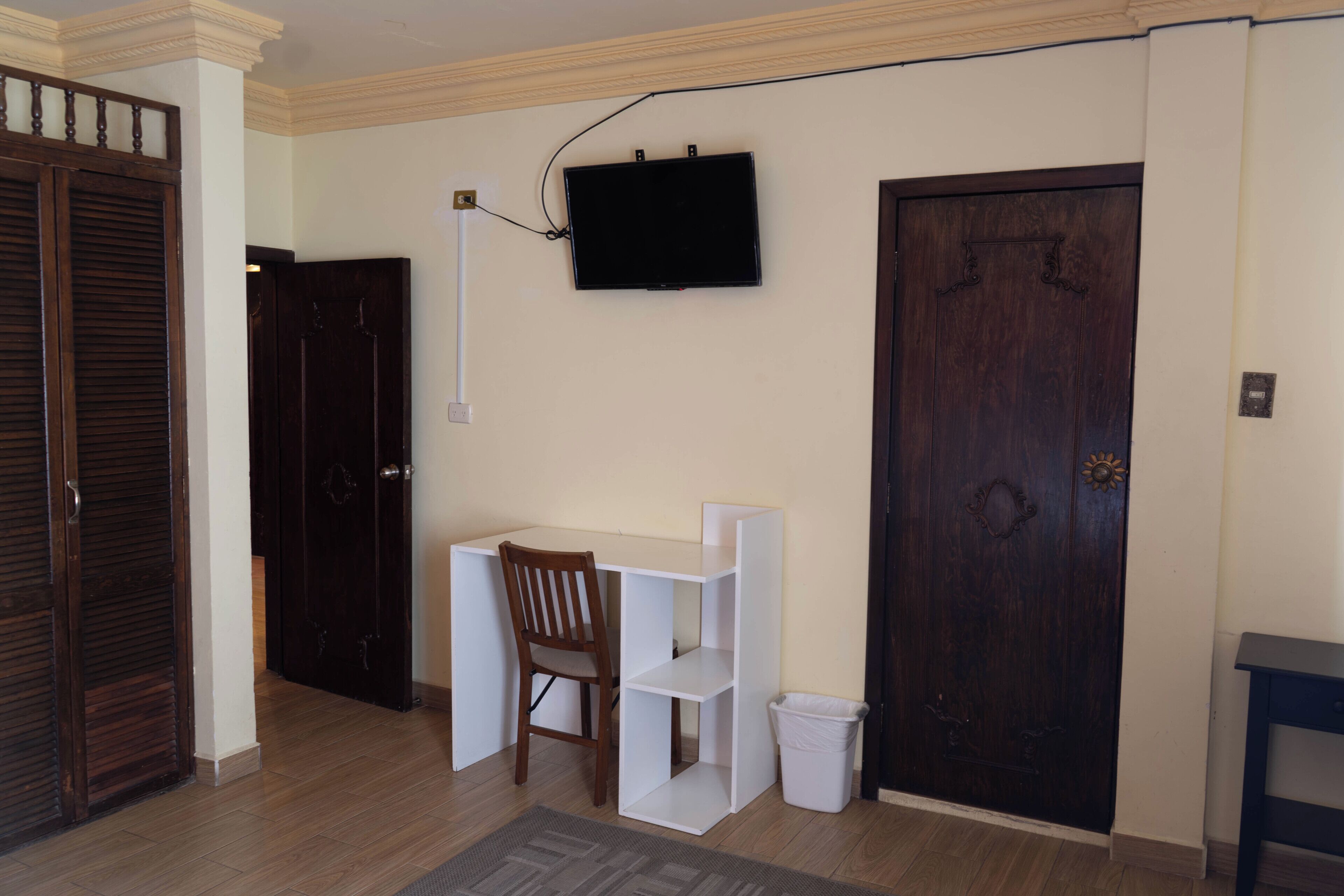 A room with a wooden desk, a chair, a television mounted on the wall, a dark wooden door, and a white trash bin.