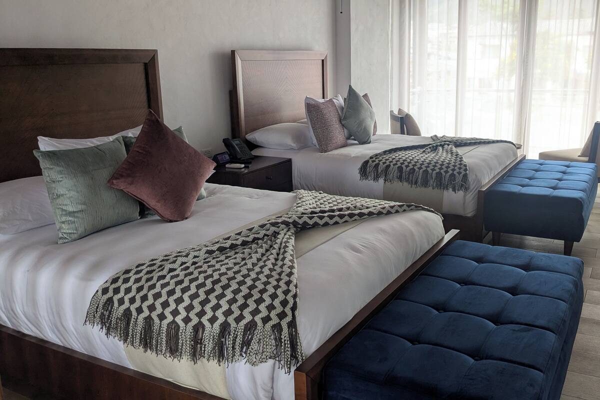 A hotel room with two beds, a bedside table with a phone, a chair, and a large window with sheer curtains.