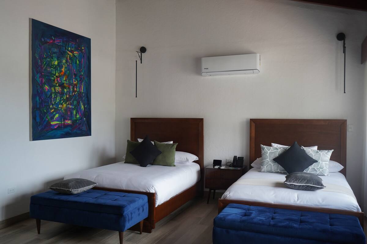 A hotel room with two beds, a painting on the wall, an air conditioner, and a small table with a phone and a lamp.