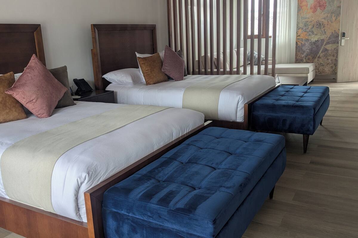 A hotel room with two beds, each with a wooden headboard, a bedside table with a phone, and a blue ottoman.