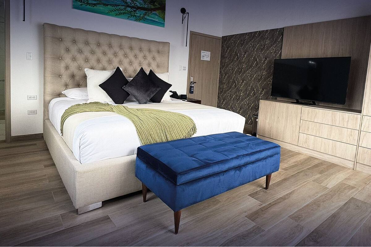 A modern bedroom with a tufted headboard, a large bed with white bedding and multiple pillows, a blue ottoman, a TV, and a marble-patterned wall.