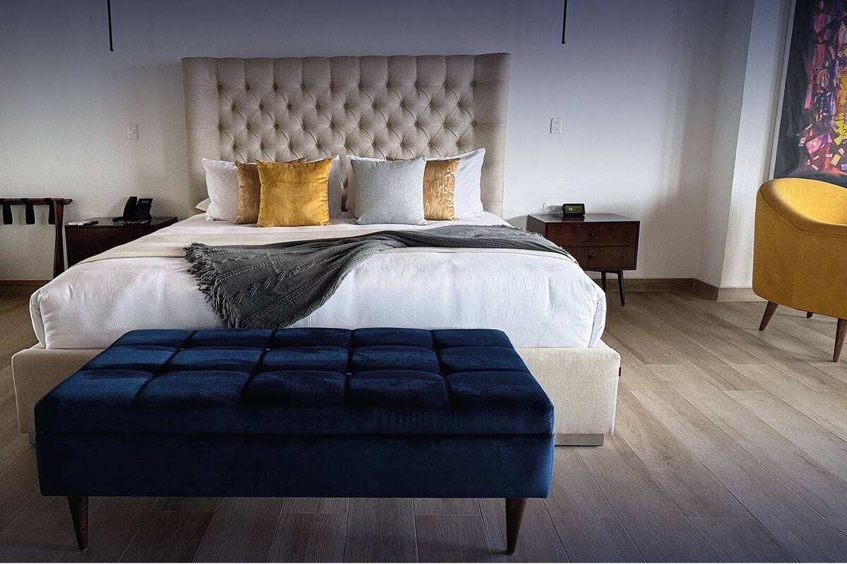 A bedroom with a large bed, a tufted headboard, multiple pillows, a bedside table with a lamp, a chair, and a bench.