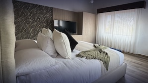 A modern bedroom with a large bed, a flat-screen TV, and a view of the outdoors through sheer curtains. - Hotel Boutique Mitad del Mundo (Pomasqui)
