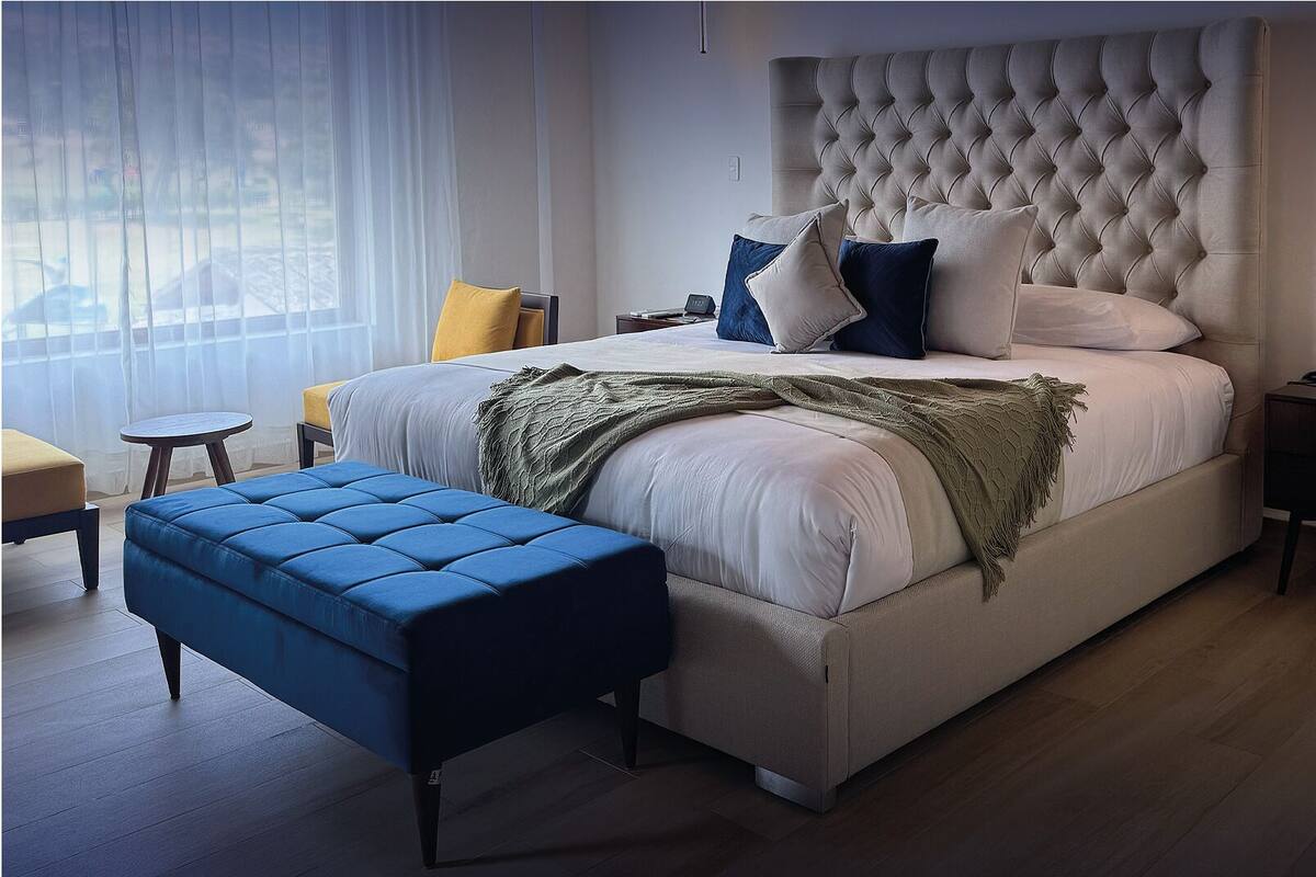 A modern bedroom with a tufted headboard, a large bed, a blue ottoman, a bedside table with a phone, and a small table with a lamp.