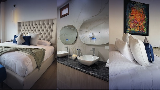 A hotel room with a tufted headboard, a large bed with multiple pillows, a bedside table, a bathroom with a vanity, a sink, and two mirrors. - Hotel Boutique Mitad del Mundo (Pomasqui)