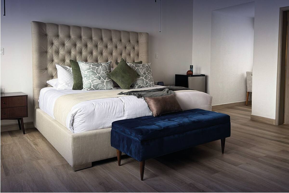 A bedroom with a tufted headboard, a bed with multiple pillows, a bedside table, a small cabinet, and a wooden floor.