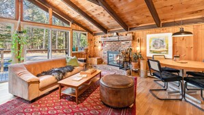 A cozy cabin interior with a wooden ceiling, a fireplace, a sofa, a dining table, and a large window overlooking a forest. - As Unique as they come! (Intervale)