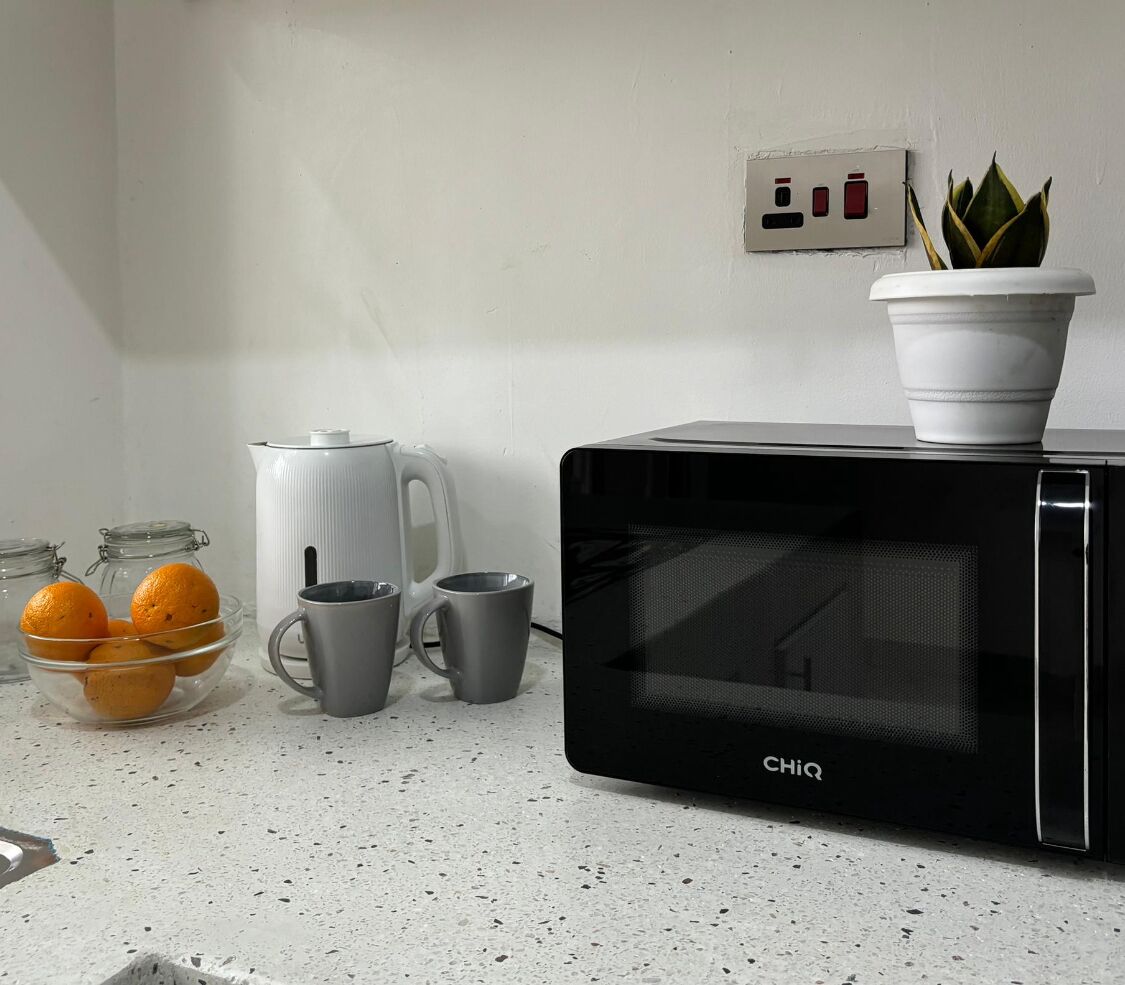 A kitchen with a black microwave, a white kettle, two grey mugs, and a bowl of oranges on the counter.