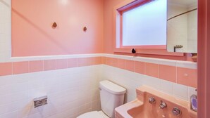A bathroom with pink and white tiles, a white toilet, a sink with a mirror, and a window above the sink. - Historic Blue Ridge Tourist Court - Queen Plus (Boone)