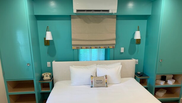 A hotel room with teal walls, a bed with white linens, a telephone on the nightstand, and a small shelf with towels. - Historic Blue Ridge Tourist Court - One Bathroom Suite (Boone)