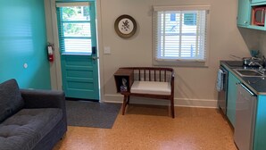 A compact living space with a teal door, a small wooden chair, a gray sofa, a kitchen area with a sink and stove, and a window with blinds. - Historic Blue Ridge Tourist Court - One Bathroom Suite (Boone)