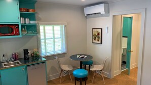 A compact kitchen with teal cabinets, a red microwave, a small refrigerator, and a window with blinds. There is a small round table with two chairs and a framed picture on the wall. - Historic Blue Ridge Tourist Court - One Bathroom Suite (Boone)