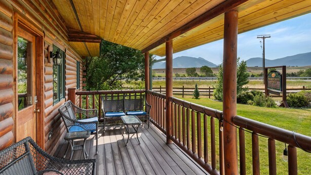 A wooden cabin with a porch featuring a table and chairs, a view of a fenced pasture, and mountains in the background. - Moose Horn Lodge- A Clean Cabin for Families, Wedding Goers & Montana Adventures (Livingston)
