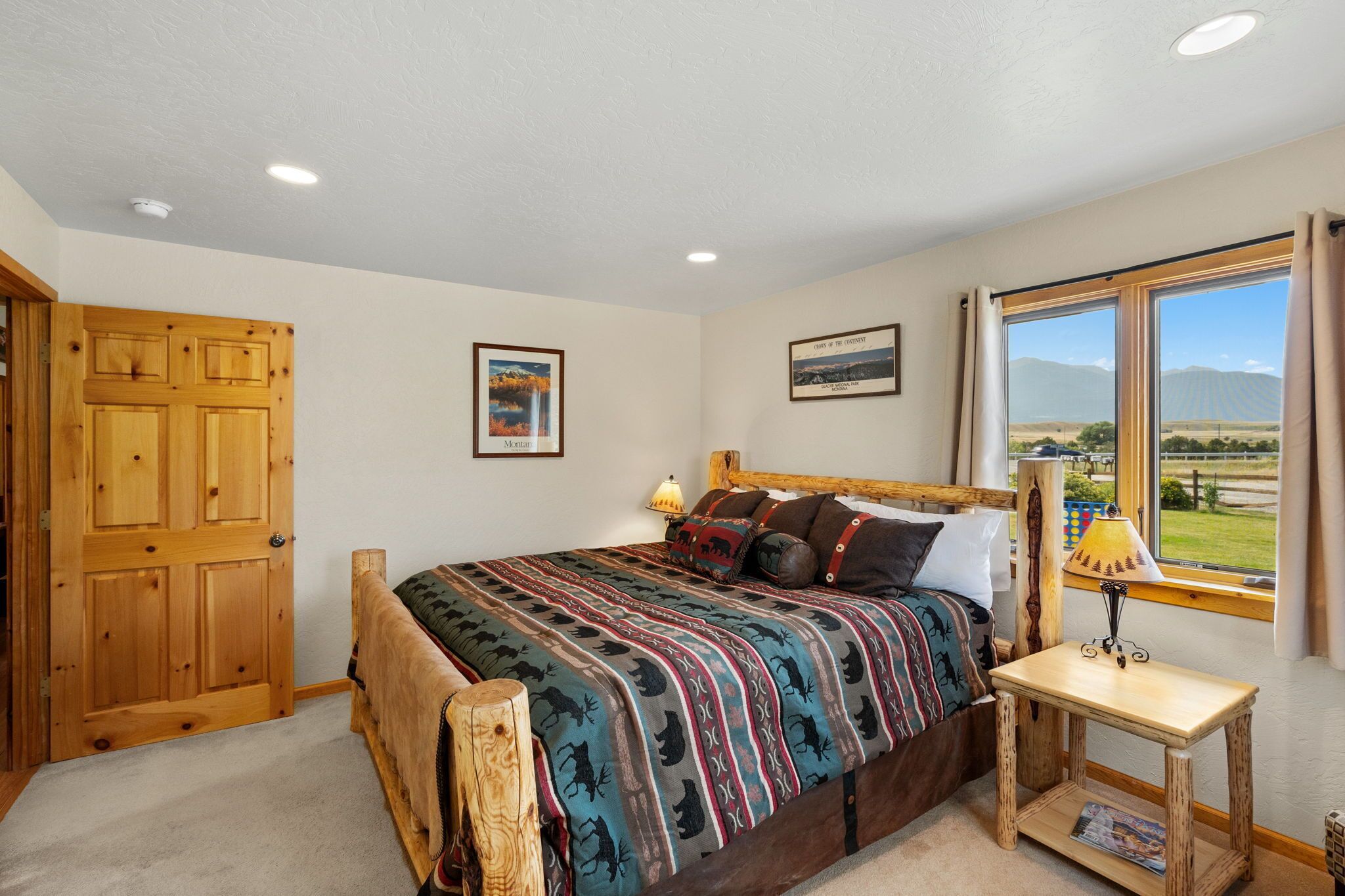 A bedroom with a wooden bed, a nightstand with a lamp, a window with a view of mountains, and a door.
