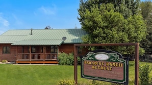 A wooden cabin with a green roof, surrounded by lush greenery and a sign that reads "Paradise Ranch Retreat". - Moose Horn Lodge- A Clean Cabin for Families, Wedding Goers & Montana Adventures (Livingston)