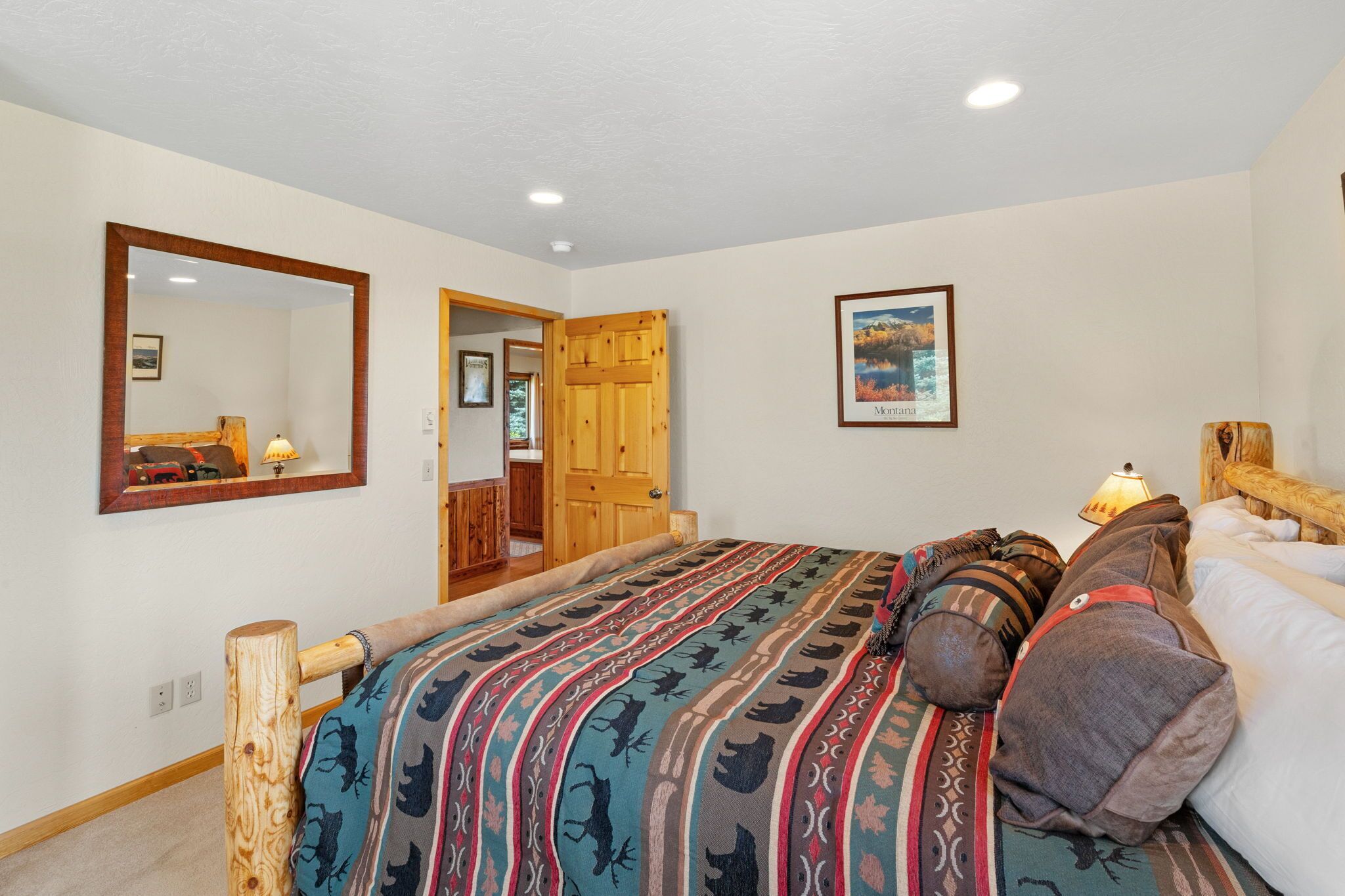 A bedroom with a large bed, a wooden headboard, a mirror, a door leading to another room, and a framed picture on the wall.