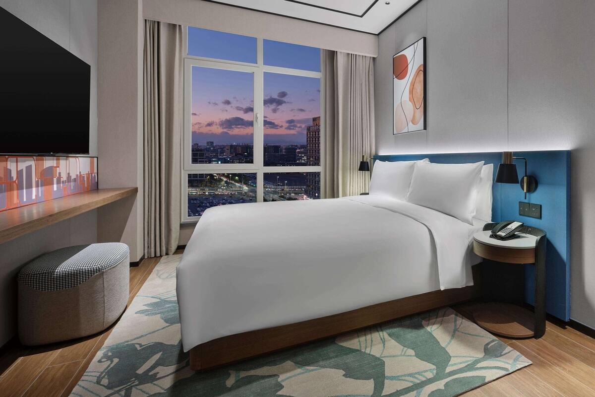 Une chambre d’hôtel moderne avec un grand lit, un téléviseur sur le côté gauche, une vue sur la ville par la fenêtre et une table de chevet avec une lampe.