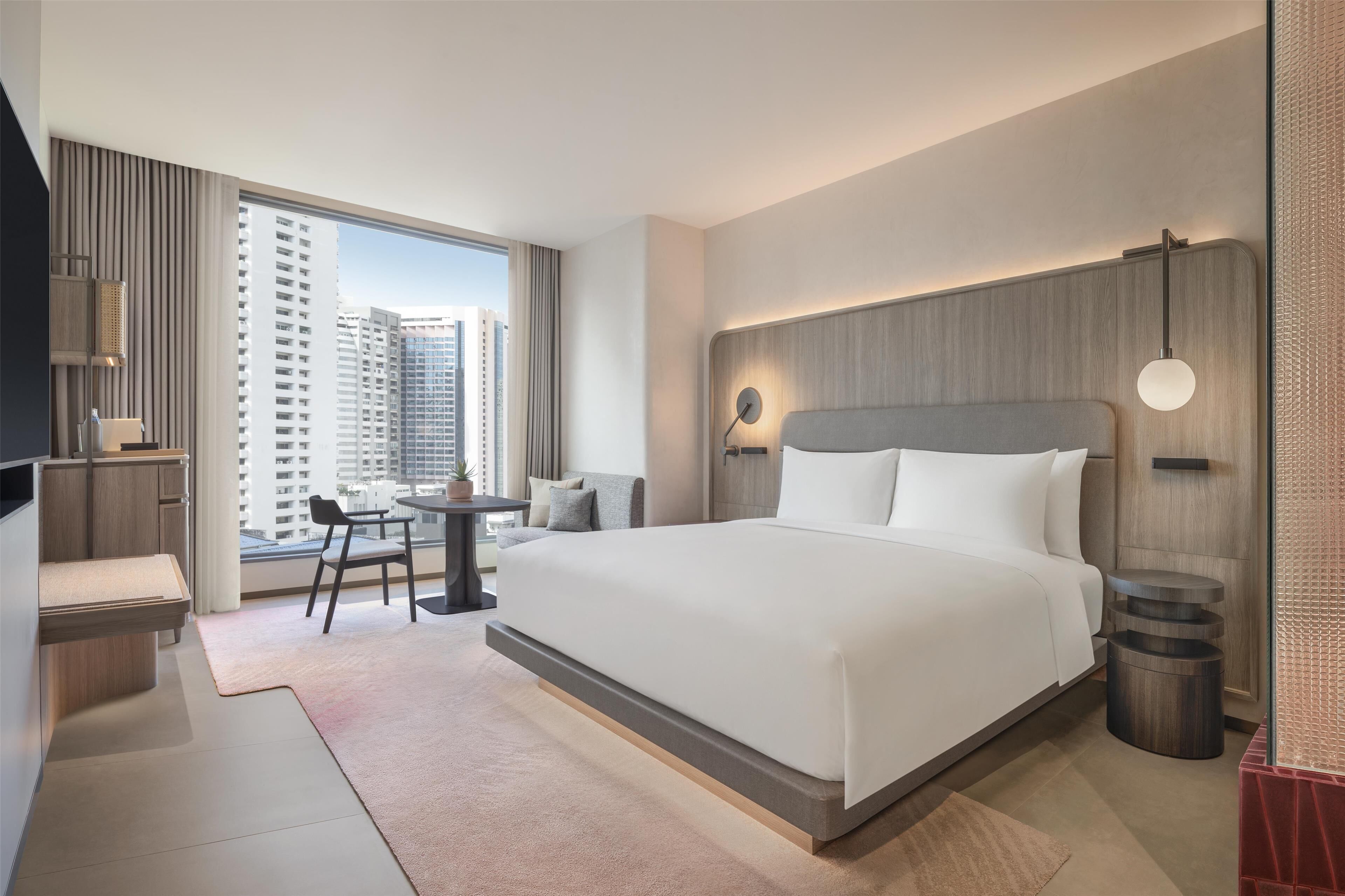 Ein modernes Hotelzimmer mit einem großen Bett, einem Schreibtisch mit Stuhl, einem Fernseher und Blick auf hohe Gebäude.