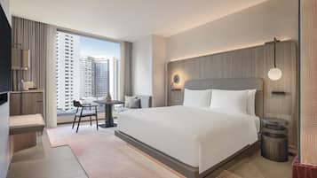 A modern hotel room with a large bed, a desk with a chair, a TV, and a view of tall buildings.