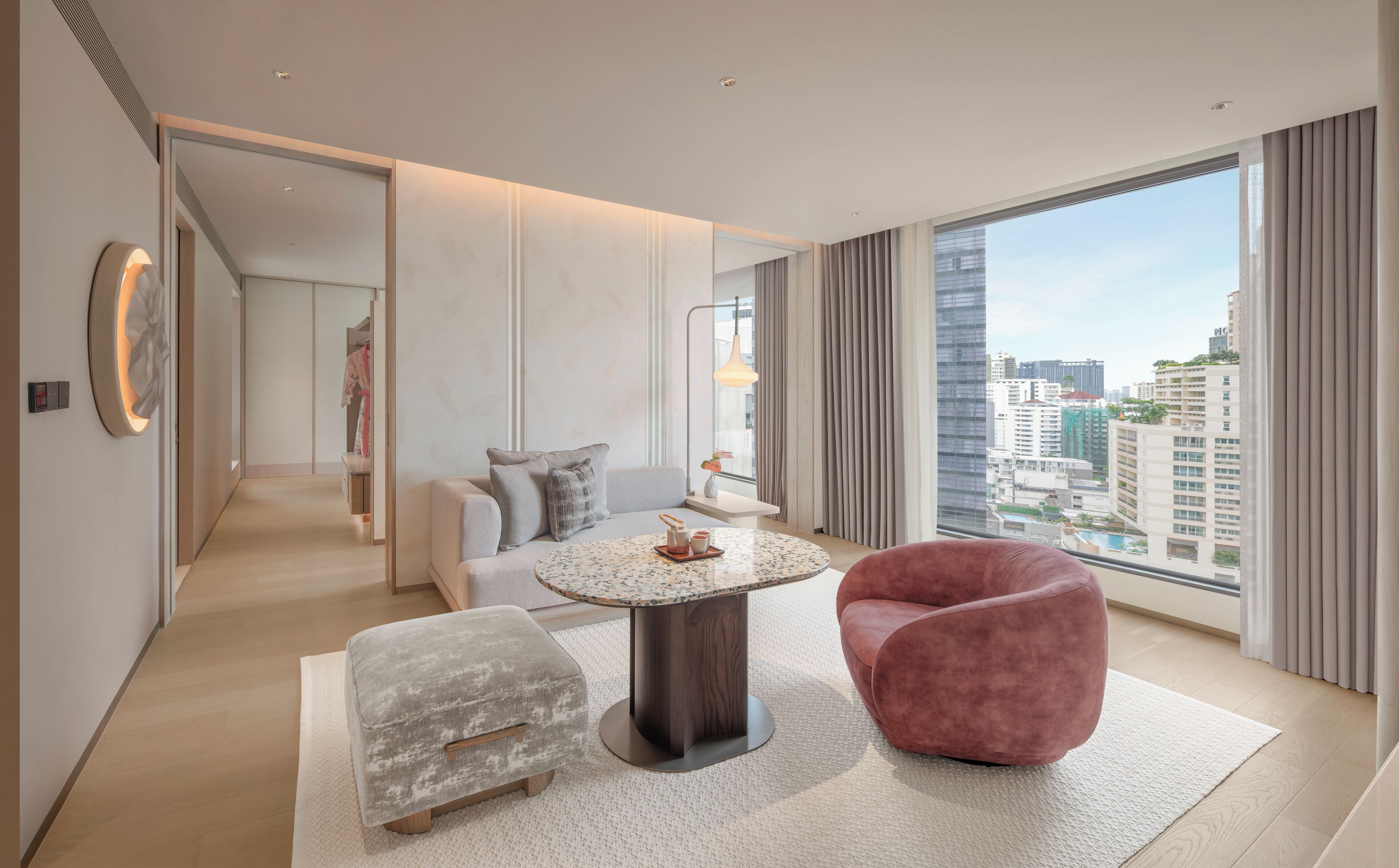 A modern hotel room with a large window, a round coffee table, a sofa, a pink chair, and a grey ottoman.