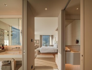 Una habitación de hotel moderna con una cama grande, baño independiente, tocador con espejo y vistas de la ciudad a través de amplias ventanas.