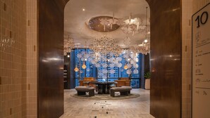 En lobby med moderne design, der fremviser et centralt siddeområde med et rundt bord og stole, en stor lysekrone og udsigt over bybilledet gennem store vinduer.