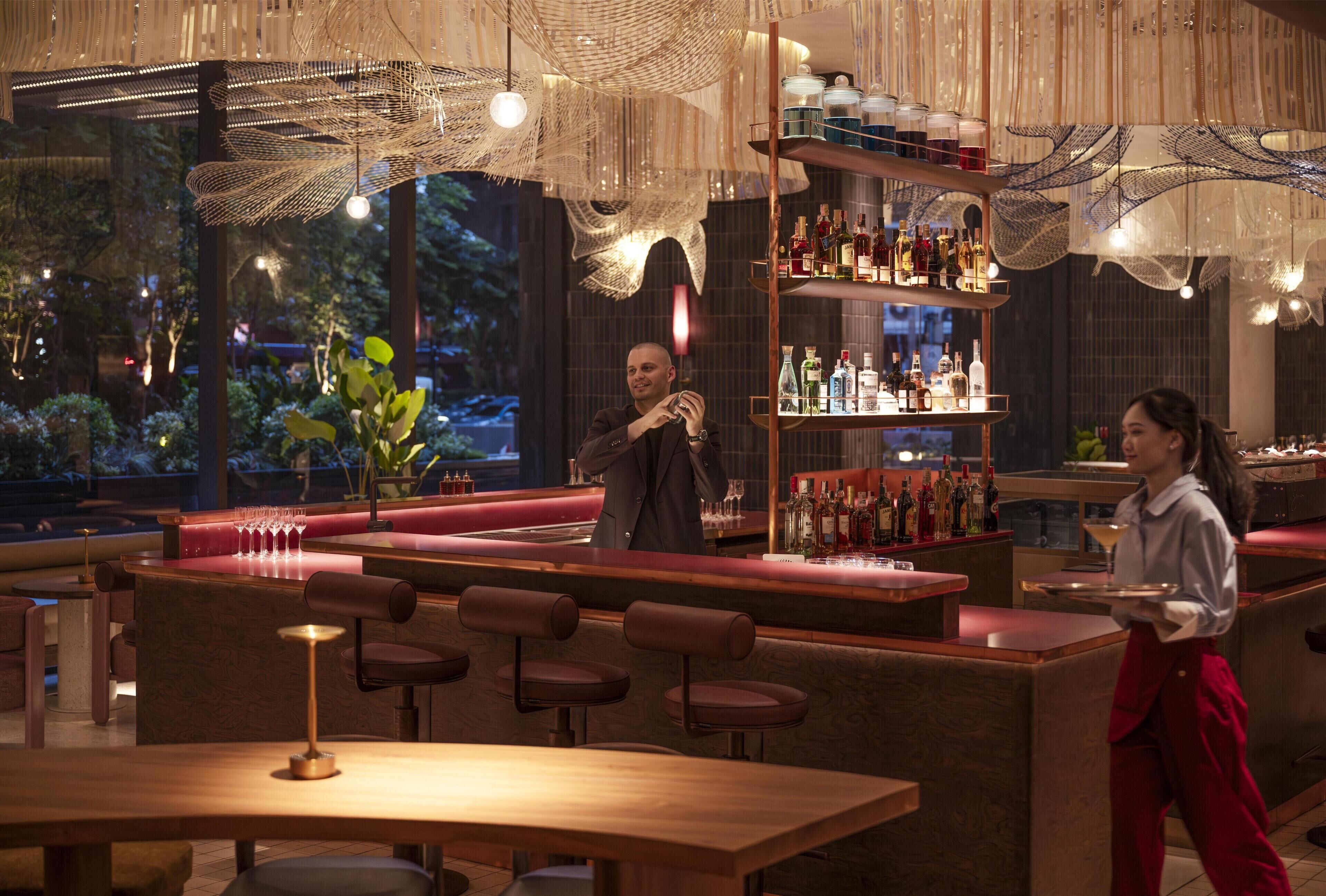 A bar area with wooden furniture, a bartender, and a well-stocked shelf with bottles.