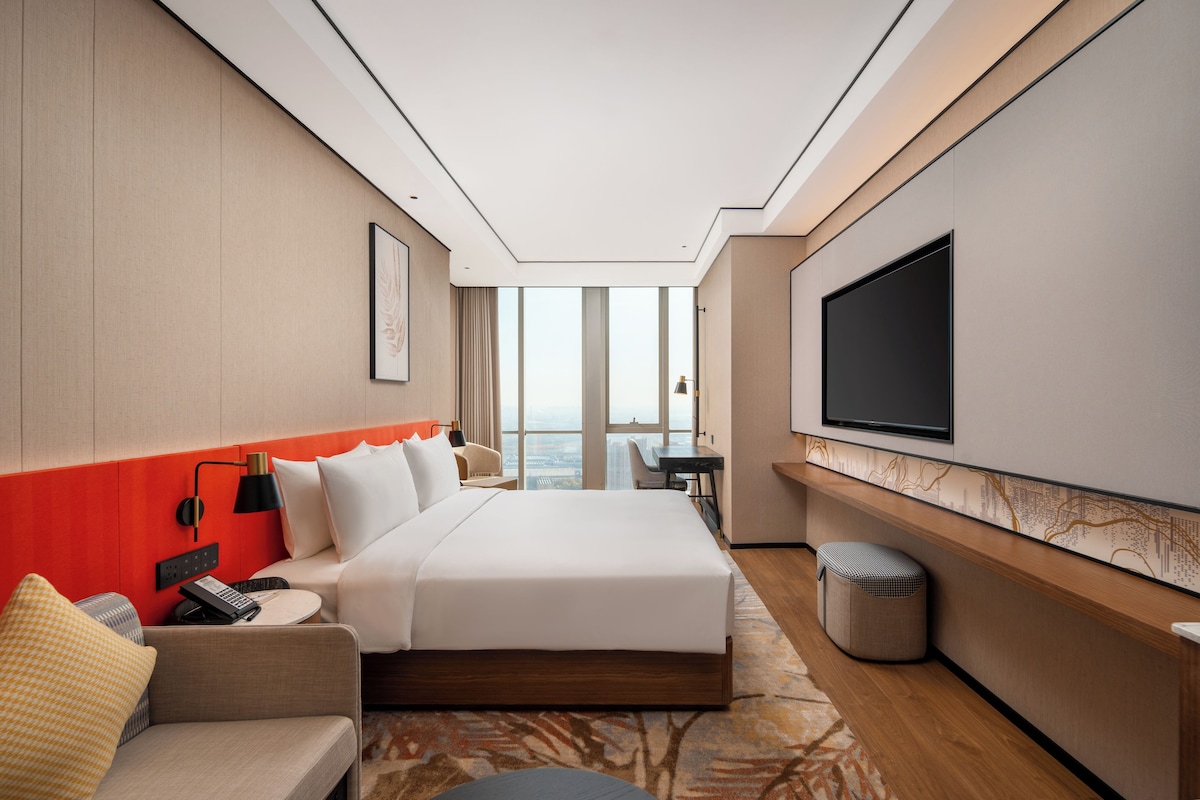 A modern hotel room with a large bed, a seating area with a sofa and armchair, a small table, a TV mounted on the wall, and a view of the city through large windows.