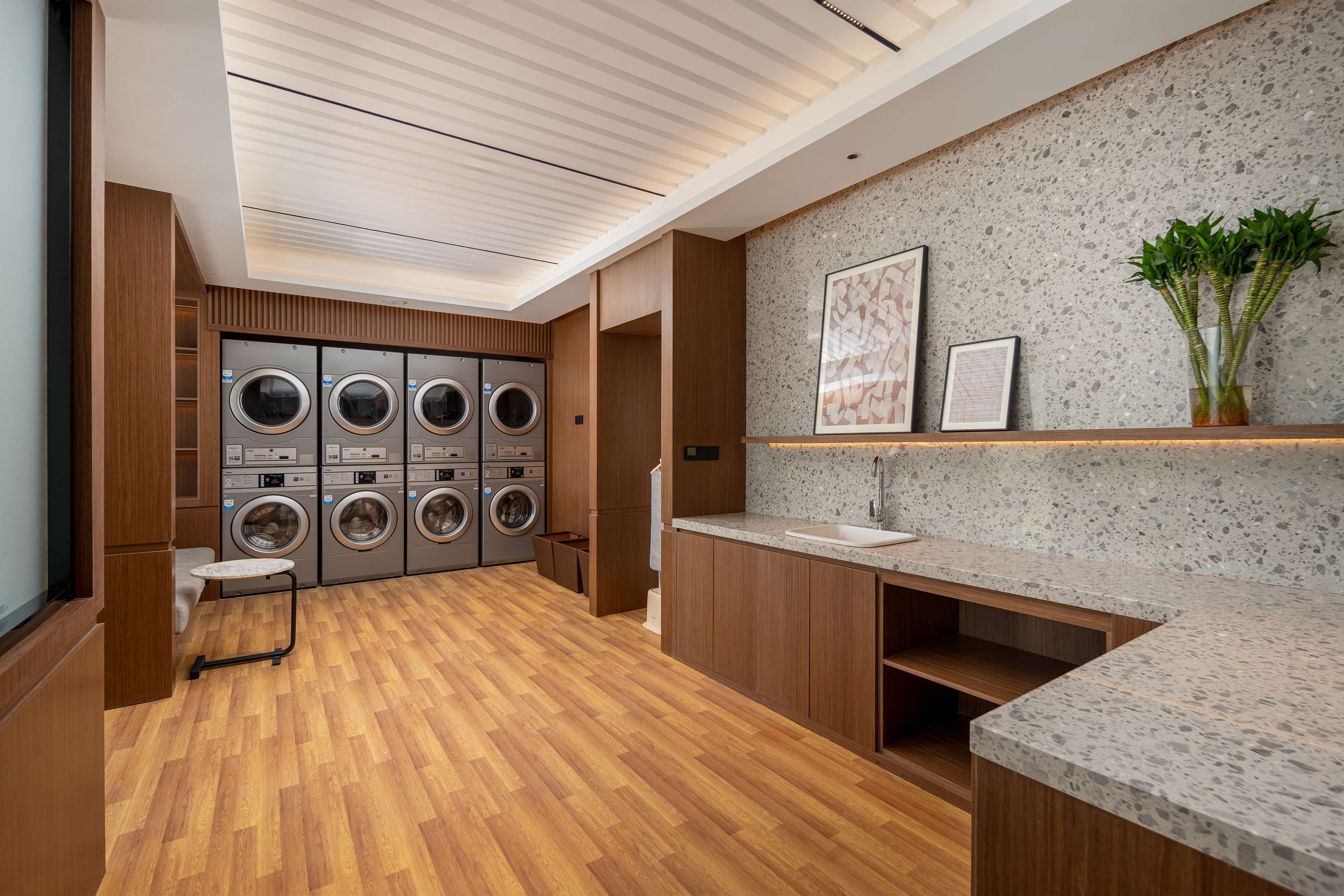A modern laundry room with multiple washing machines, wooden flooring, a sink, and a speckled backsplash.