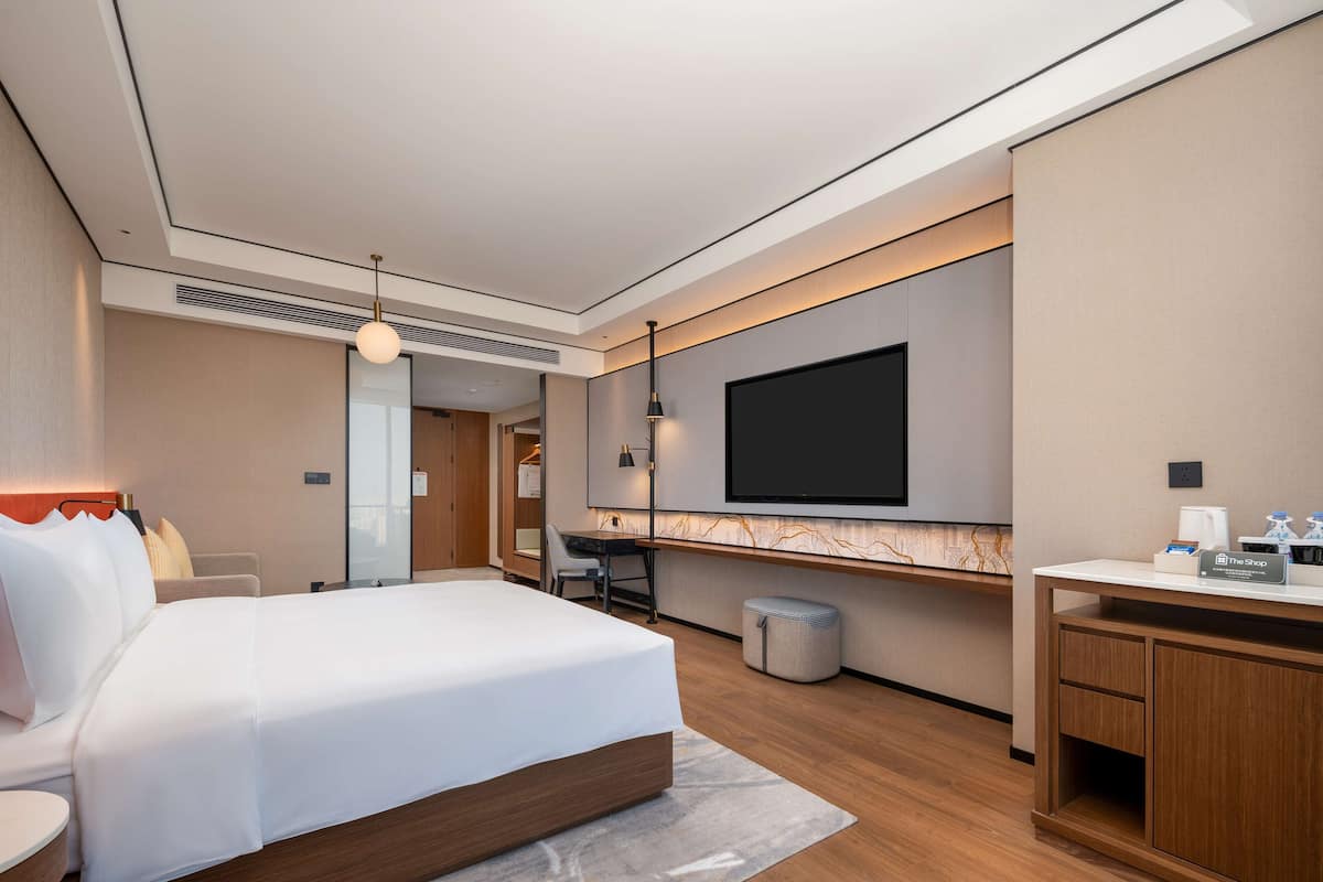 A modern hotel room with a large bed, a desk, a flat-screen TV, and a minibar.