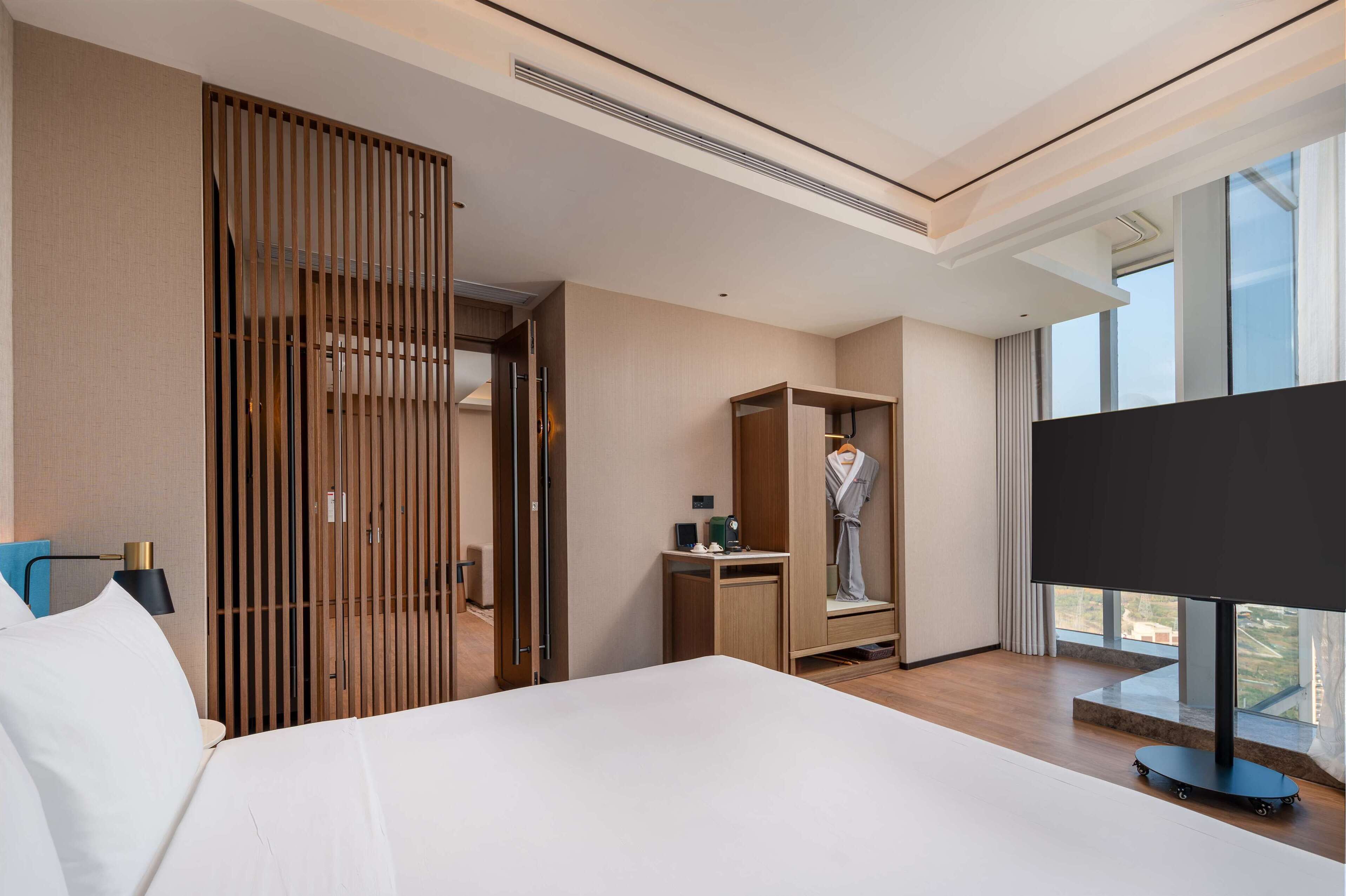 A modern hotel room with a large bed, a wardrobe, a desk, and a TV.