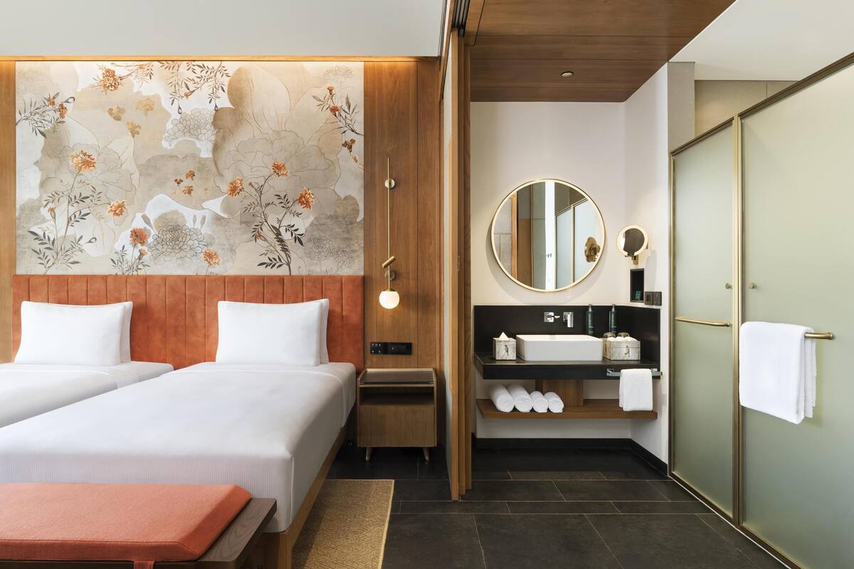 Une chambre d’hôtel avec deux lits, une tête de lit en bois, une grande fresque murale à motifs floraux, un miroir rond, un lavabo et un dressing.