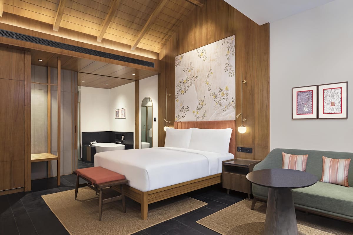 Une chambre d’hôtel moderne avec un plafond en bois, un grand lit avec du linge de lit blanc, un canapé vert, une petite table ronde et un banc en bois.