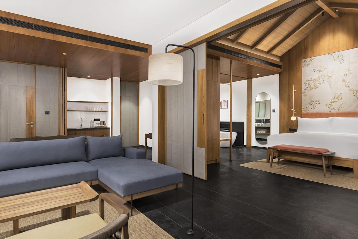 Une chambre d’hôtel moderne comprenant un canapé d’angle, une petite table en bois, un banc, un grand lit et une salle de bain séparée.
