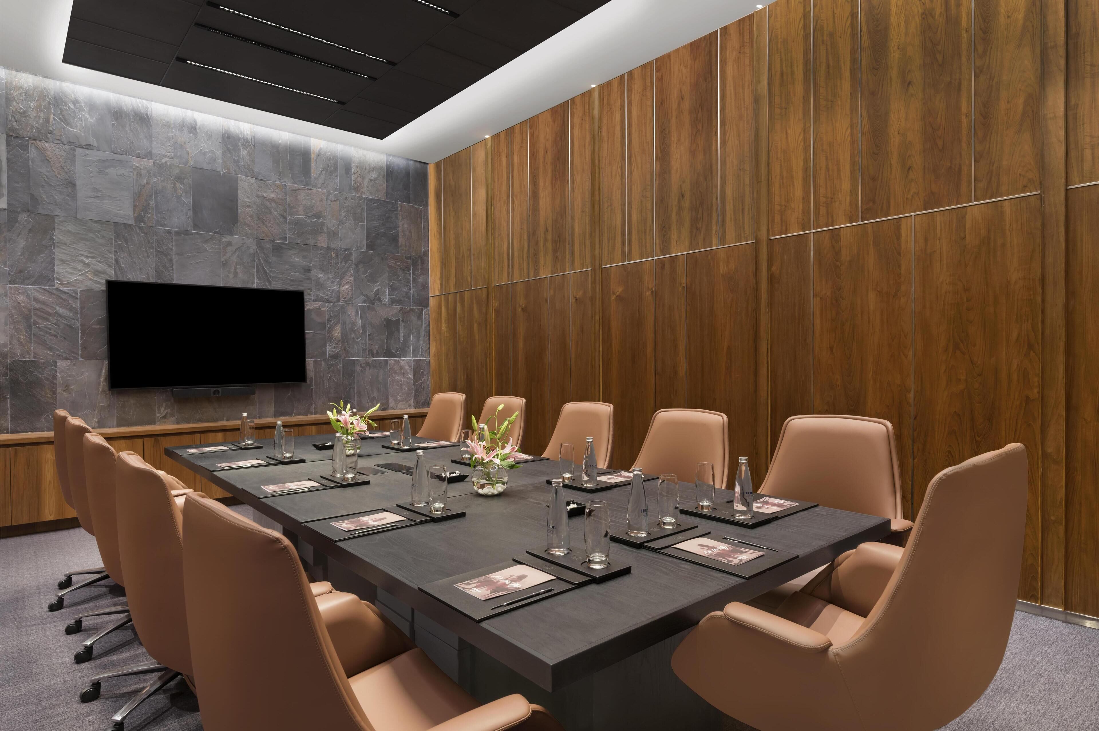 A conference room with a long table, leather chairs, a flat-screen TV, and a stone wall.