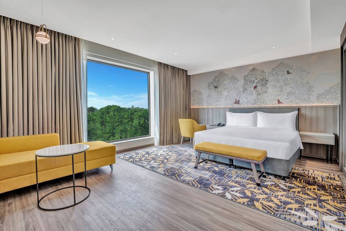 A modern hotel room with a large window, a bed, a yellow armchair, a small table, and a patterned rug.