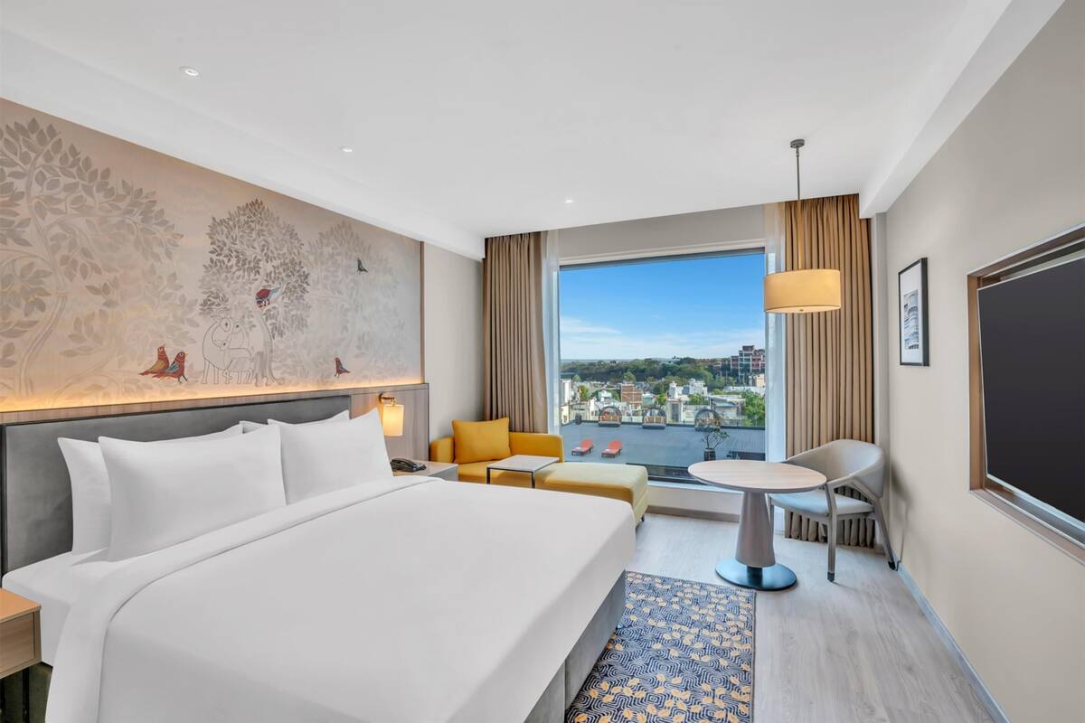 A hotel room with a large bed, a small round table, a sofa, and a view of the city through a large window.