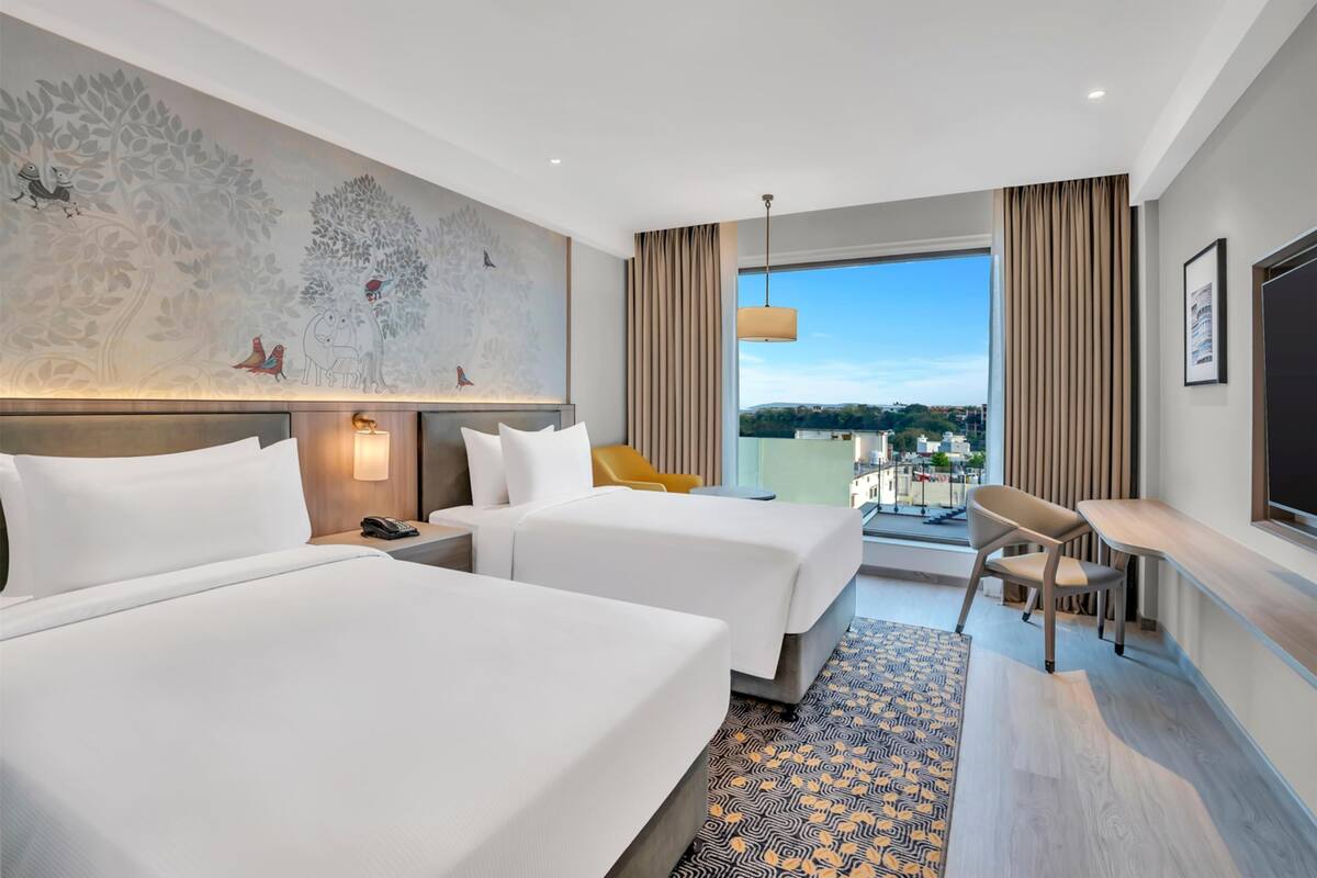A hotel room with two beds, a large mural, a view of the city, and a balcony with seating.