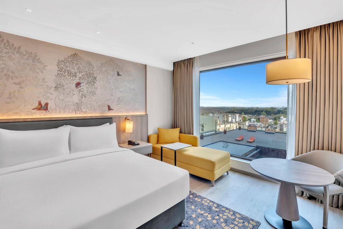 A hotel room with a large bed, a sofa, a small table, and a view of the city.