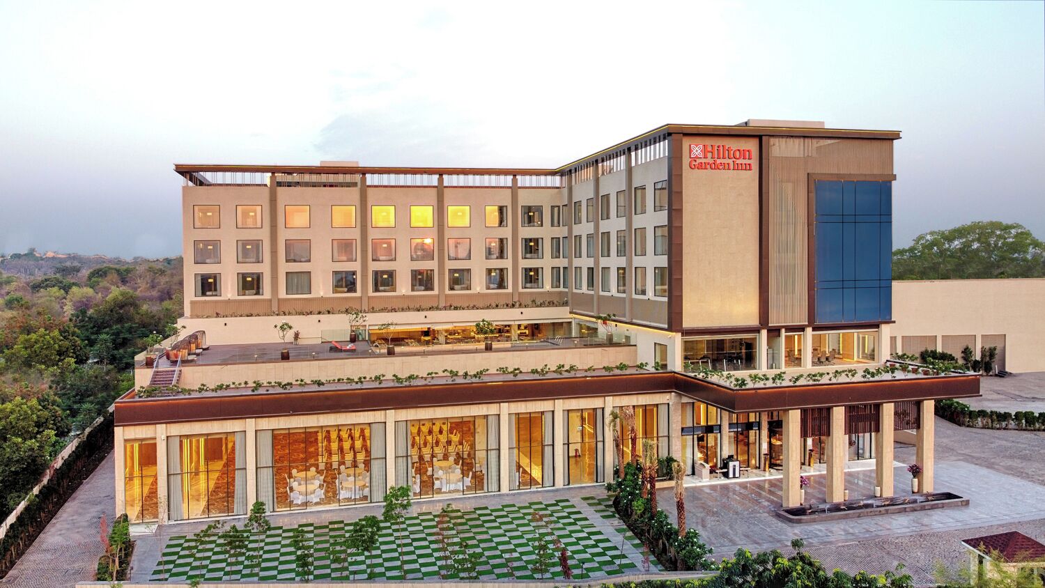 Un hotel moderno con una amplia fachada de vidrio, un jardín con diseño a cuadros y un llamativo letrero de Hilton Garden Inn.