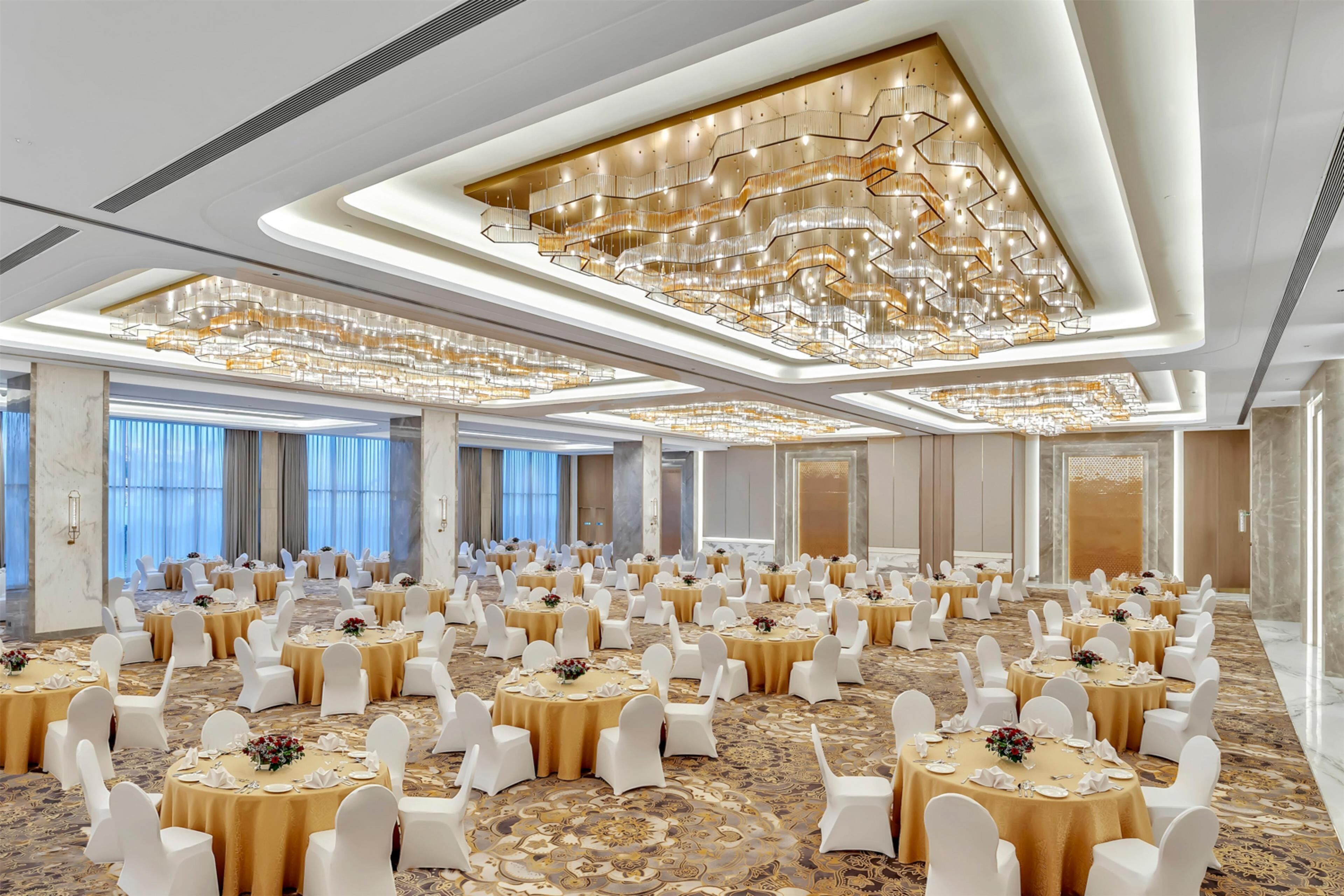 A banquet hall with multiple round tables, white chairs, and gold tablecloths, featuring a modern chandelier and large windows.