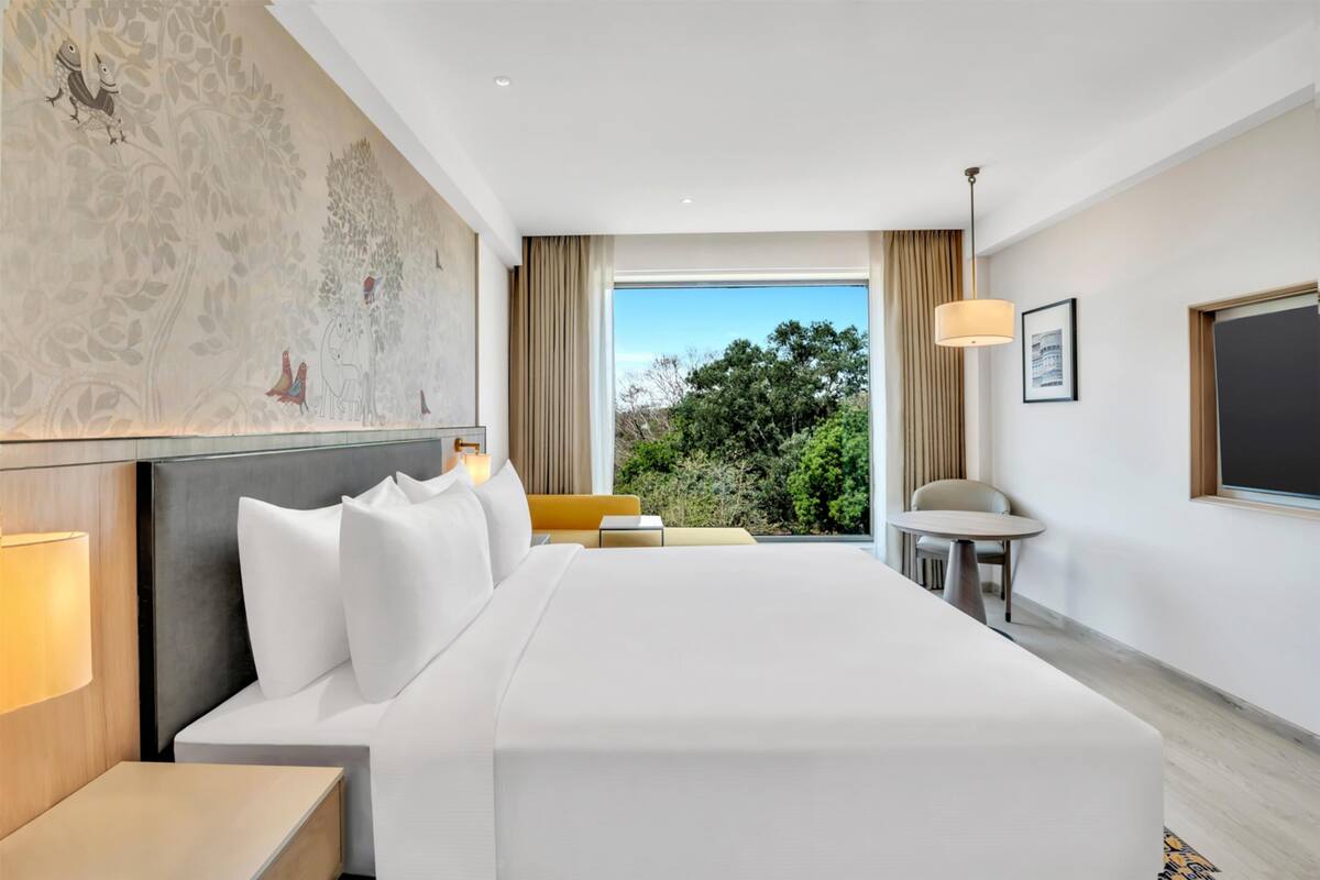 A hotel room with a large bed, a small sofa, a table, and a view of trees through the window.