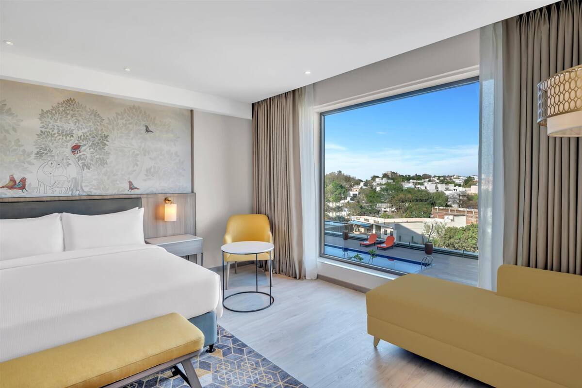 A modern hotel room with a large bed, a sofa, a small table, and a chair. There is a large window with a view of buildings and trees.