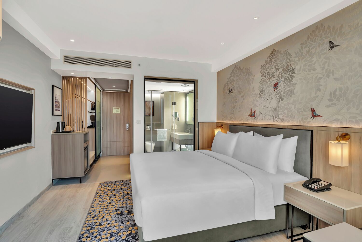 A modern hotel room with a large bed, bedside table, and a desk with a lamp.
