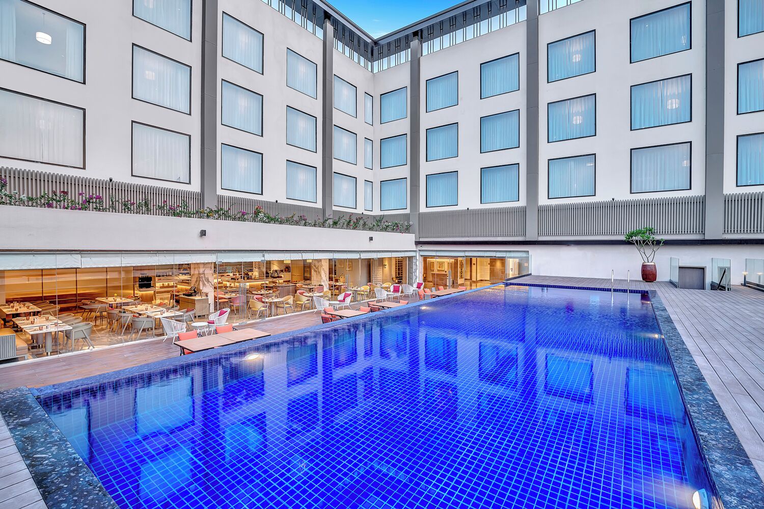 A modern hotel with a central swimming pool, outdoor seating area, and a restaurant.