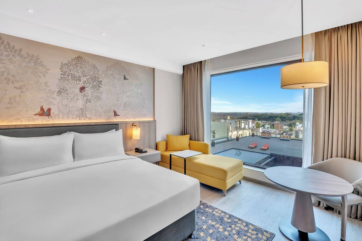 A hotel room with a large bed, a sofa, a small table, and a view of the city through a large window.